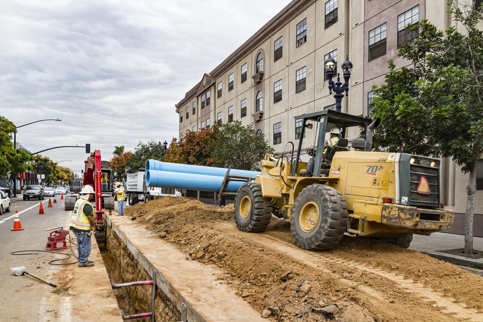 13-027 Sewer and Water Group 957, machinery and crew excavating deep trench and laying water pipe in busy commercial district